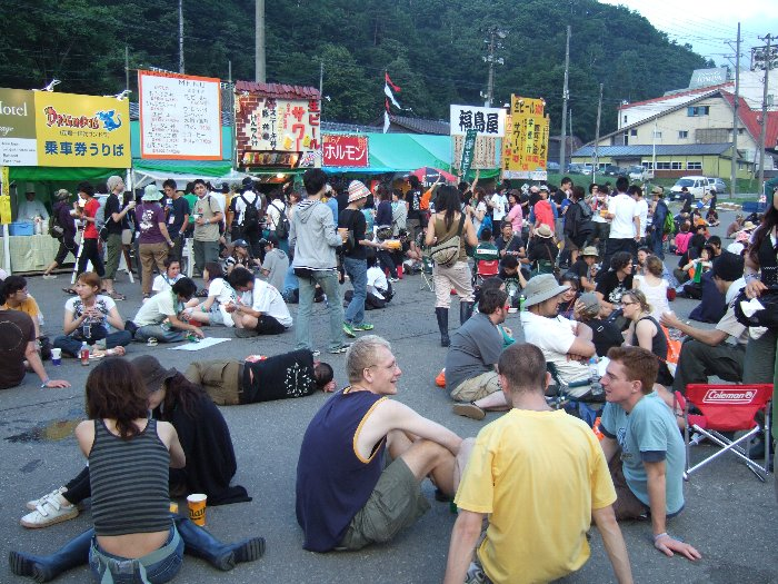 場外ショップエリアの飲食店前。前夜祭の午後6時ごろ。いすやテーブルはありませんが、地面がアスファルトなのでそのまま座って食べる人が多い。